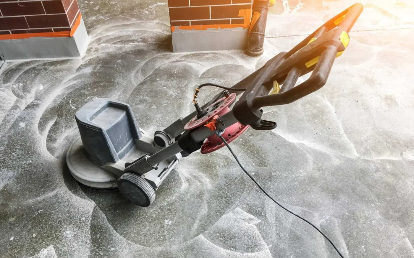 A floor cleaning machine is being used on a concrete floor, leaving circular cleaning patterns. The machine has a grey body and black handle, with wires extending from it. The background shows brick walls and sunlight filtering through. For professional results like this in Gardnerville NV, contact our concrete contractors for a free quote.