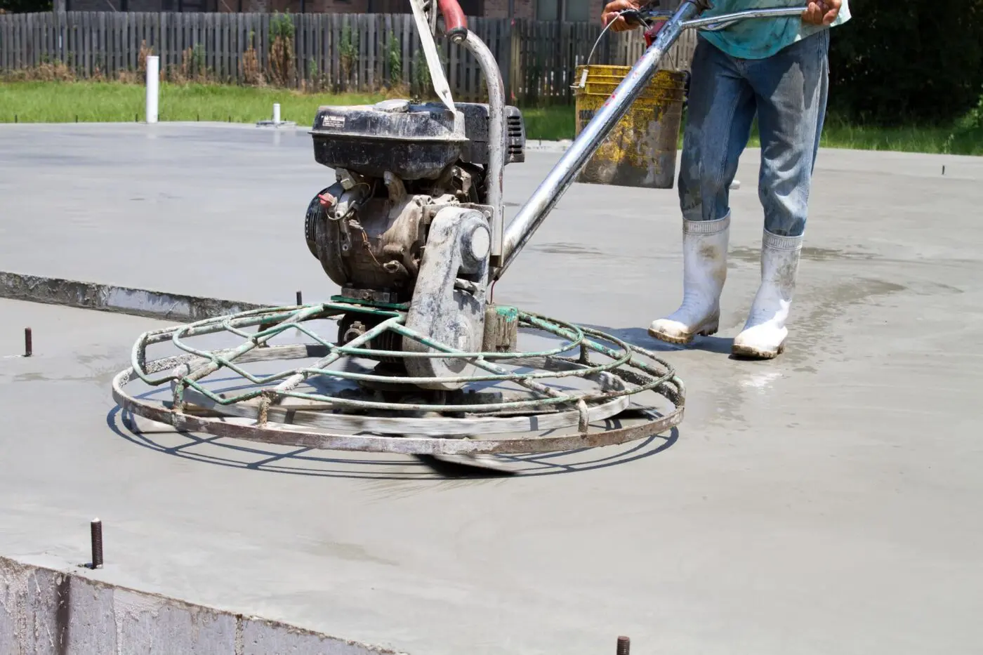 A worker in white rubber boots and jeans operates a power trowel to smooth the surface of a large concrete slab, ensuring all concrete needs are met. The worker holds onto the machine's handle with its rotating metal cage while standing on the freshly poured concrete. A fence is visible in the background.