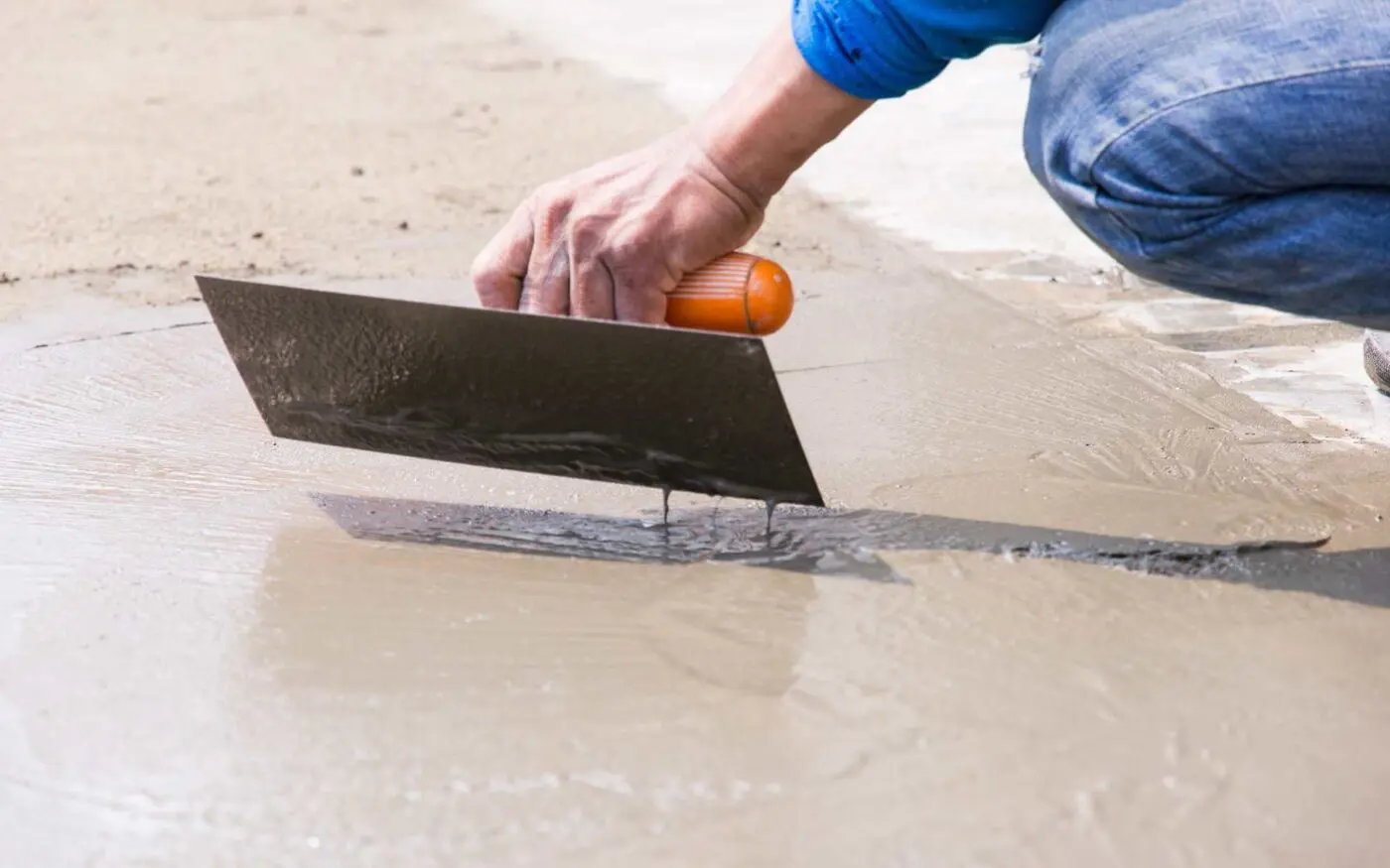 A close-up of a person using a trowel to smooth out a freshly laid cement surface. The worker, wearing a blue shirt, is in a kneeling position and firmly holding the trowel with an orange handle, ensuring an even and smooth finish on the wet cement—showcasing Reno Concrete Solutions' quality concrete work in NV.