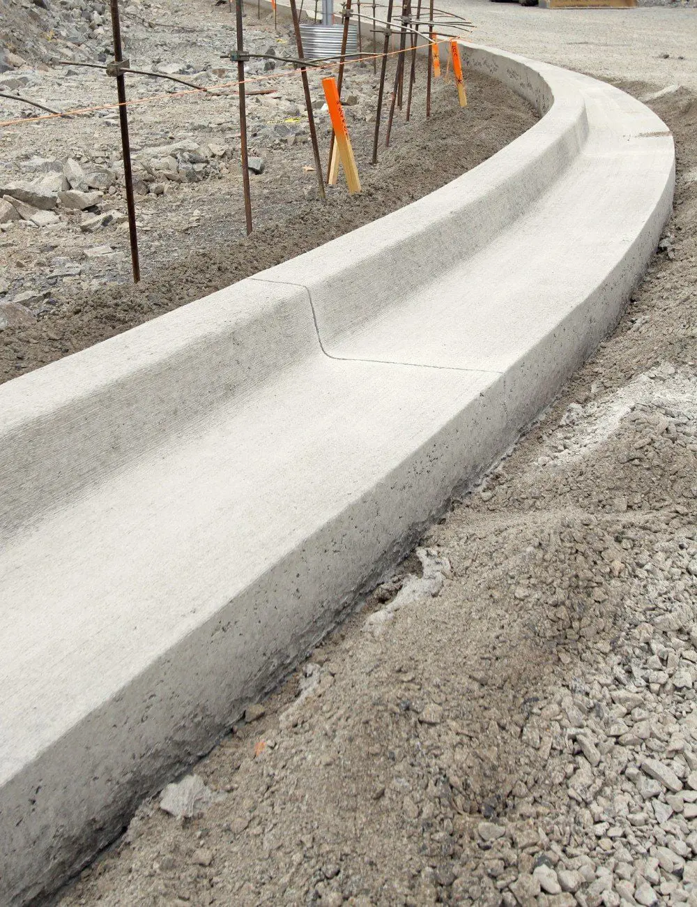 At a construction site, Reno Concrete Solutions has constructed a slightly curved concrete curb, surrounded by gravel, dirt, and rebar. Orange markers are visible on metal rods in the background. For details or a free quote on your next project, including Sun Valley decorative concrete options, contact us today.