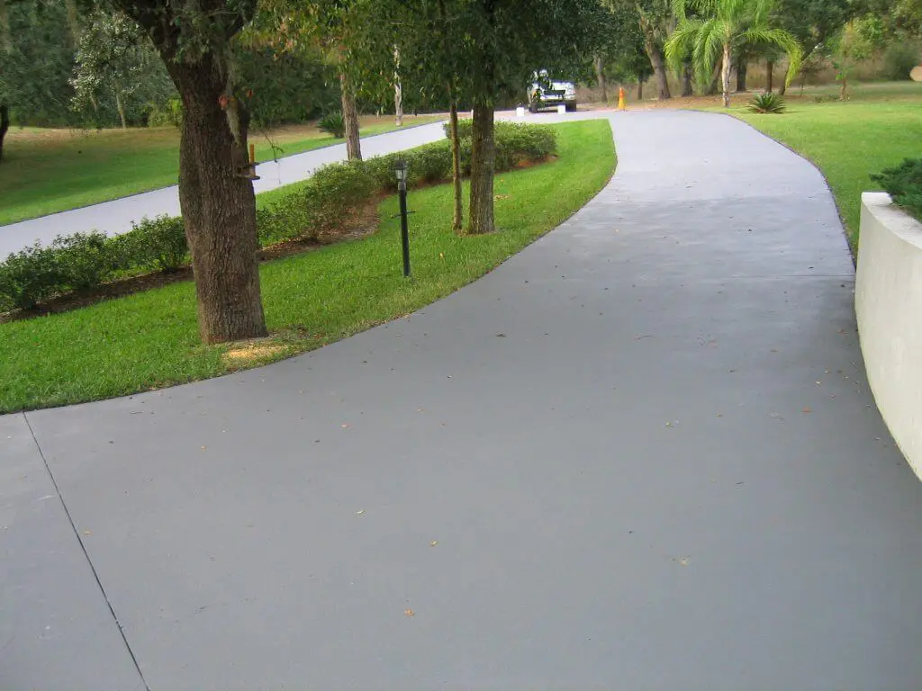A smooth, gray concrete driveway curves around a landscaped yard with well-maintained grass, shrubs, and trees. In the background, a golf cart is visible approaching the curve of the driveway. The setting appears peaceful and shaded, ideal for showcasing top-notch resurfacing services available in Reno NV.