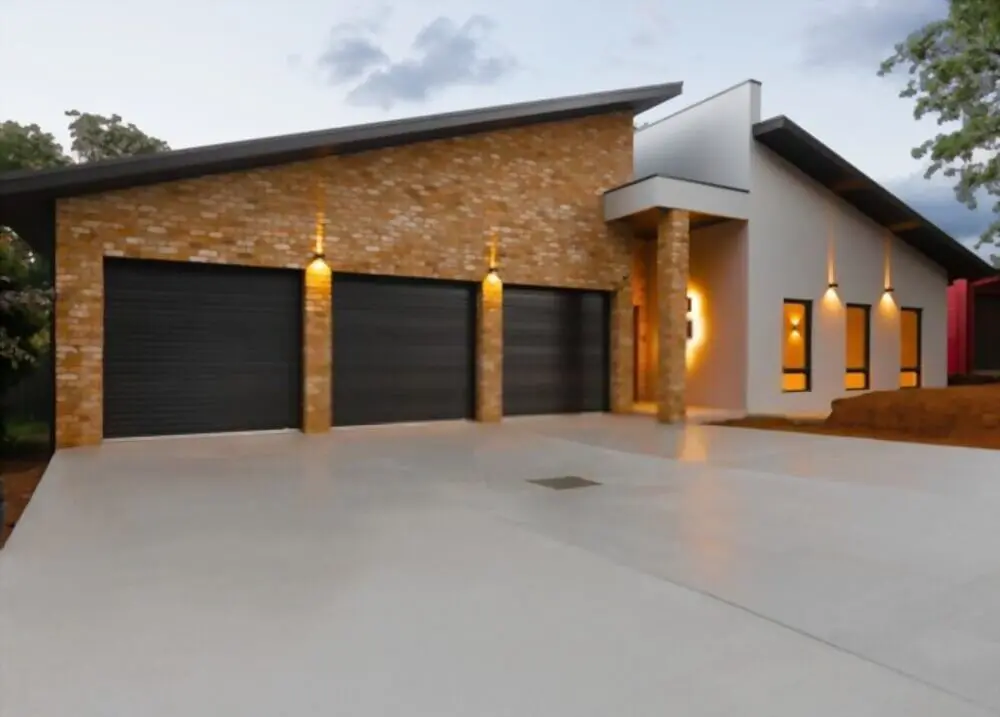 A modern single-story house in Gardnerville, NV with a sloped roof, featuring a brick facade and a white exterior. The house has a large driveway leading to four black garage doors. Licensed concrete contractors created the stunning driveway. Warm lights illuminate the brick pillars at dusk.