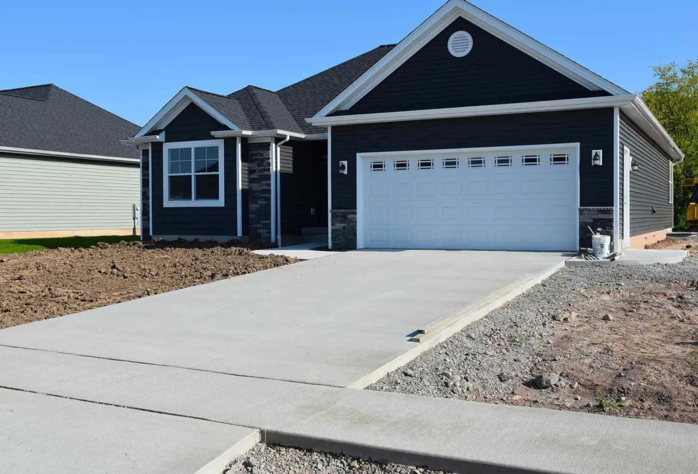 A newly built modern house with dark gray siding and a white garage door. The concrete driveway and sidewalk, thanks to Reno Concrete Solutions, are freshly laid, contrasting the bare dirt yard where landscaping is yet to be done. Under the clear blue sky of Genoa NV, this home awaits its finishing touches.