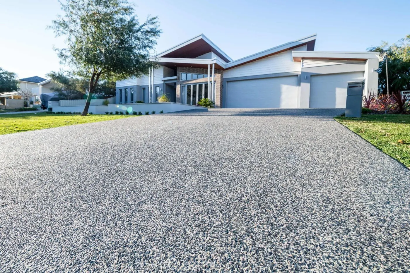Modern single-story house with a wide driveway leading up to a three-car garage. Featuring Sun Valley decorative concrete by Reno Concrete Solutions, the sleek design boasts large windows and a landscaped front yard with green grass and trees. The sky is clear and blue.