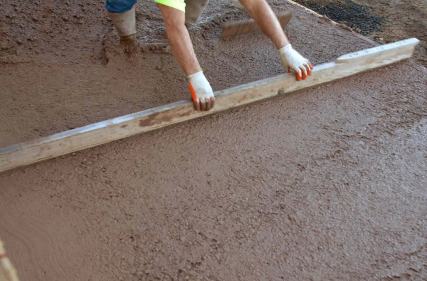 A person in white gloves spreading concrete with a straightedge tool on a construction site, representing Reno Concrete Solutions. The individual, wearing a yellow shirt and partly visible boots, ensures the surface remains smooth after being worked on for quality concrete solutions.