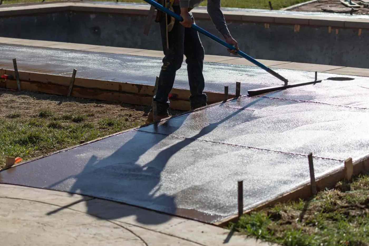 A licensed concrete contractor smooths the surface of freshly poured concrete with a long-handled tool. The concrete sections are framed with wooden boards and metal stakes, and there is a swimming pool in the background. The sun casts the worker’s shadow on the concrete, Gardnerville NV. Contact for a free quote.