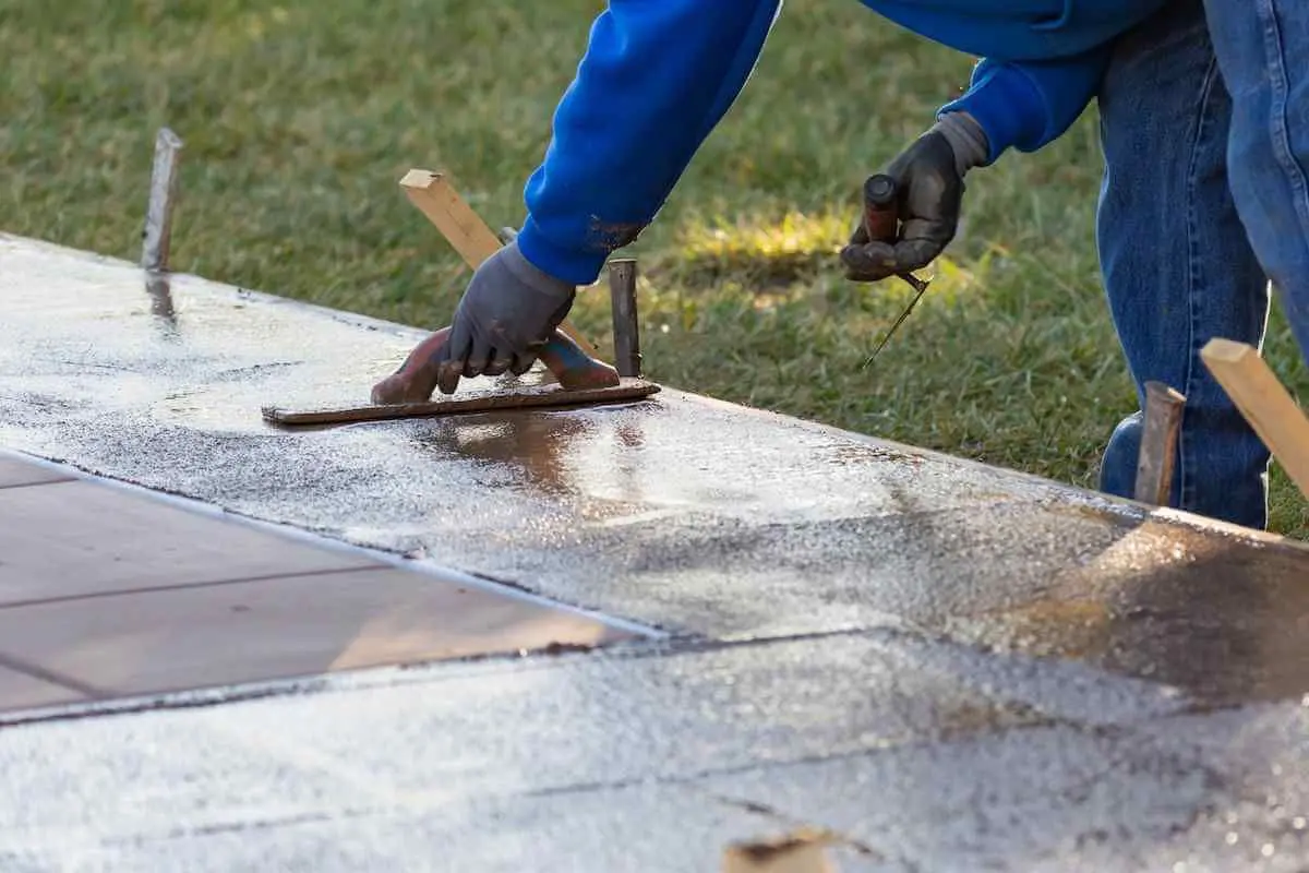 A person in a blue hoodie and gloves uses a trowel to smooth out freshly poured concrete, ensuring quality concrete solutions. The concrete is sectioned off with wooden stakes, and grass can be seen in the background.