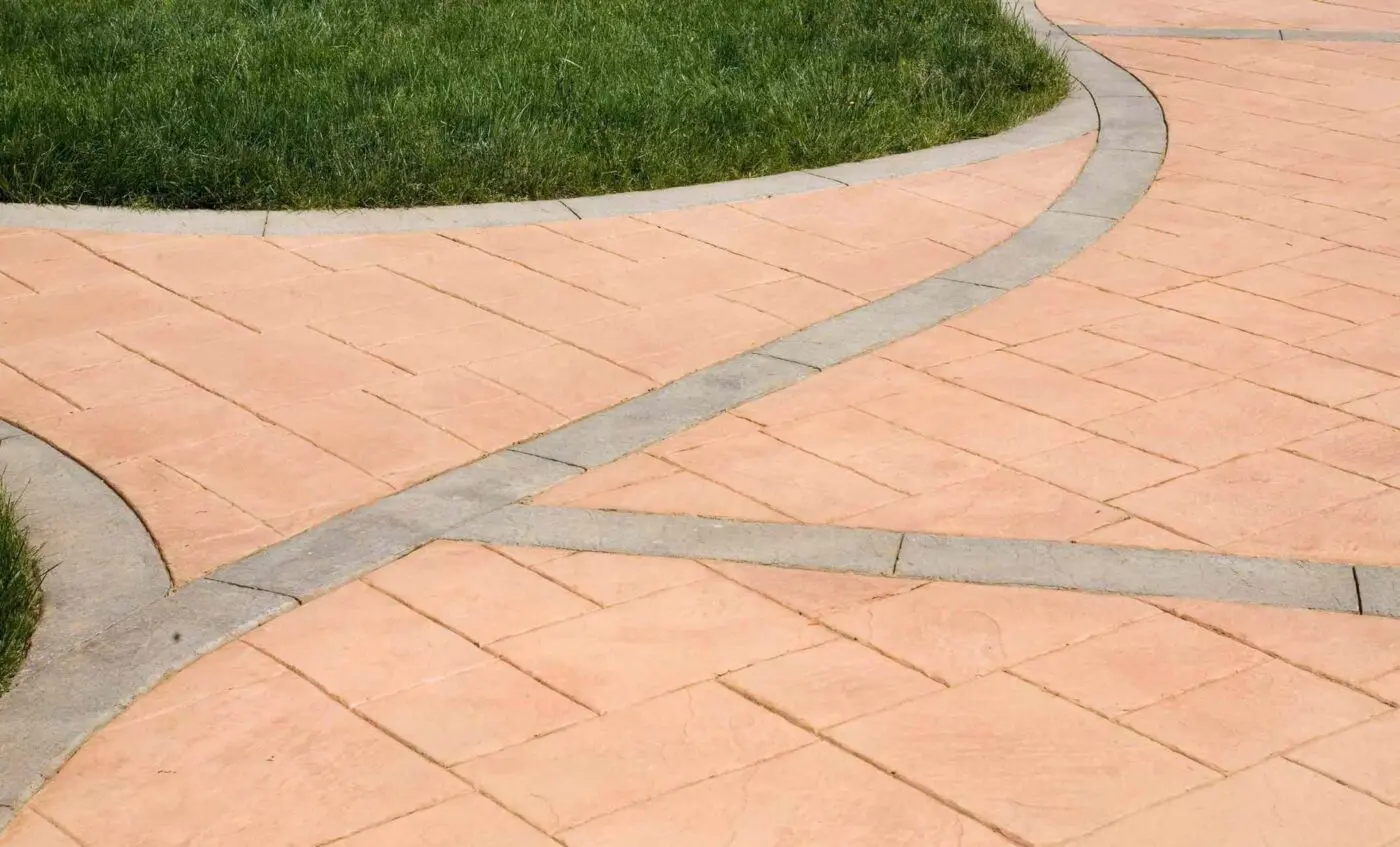 A paved, salmon-colored concrete walkway by Reno Concrete Solutions features intersecting paths with beige borders, winding through a grassy green lawn. The neat geometric design adds structure and visual interest to the landscaped area, showcasing the quality concrete solutions provided by this team.