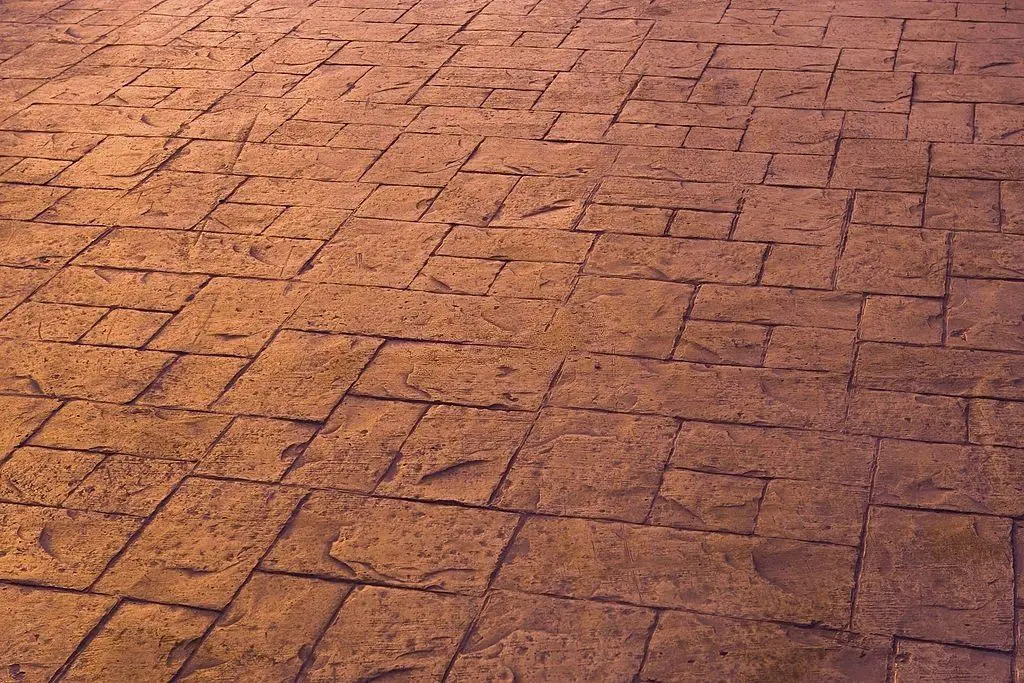 A textured, stone-paved surface with an irregular grid pattern. The stones vary in size and have a weathered, reddish-brown appearance, with visible lines and grooves creating a rustic look. Get this unique design from Reno Concrete Solutions. Contact them today for a free quote!