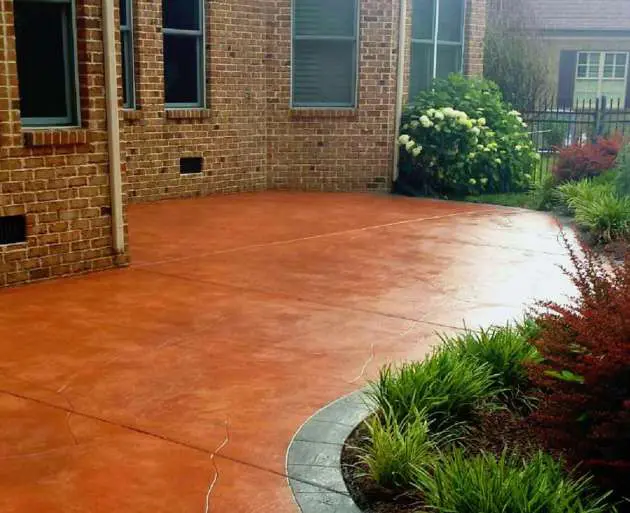 A backyard patio with terracotta-colored concrete flooring, expertly crafted by Reno Concrete Solutions, is bordered by green plants and shrubs. Adjacent to a brick house with multiple windows, the patio overlooks a garden area abundant with greenery in Genoa NV.
