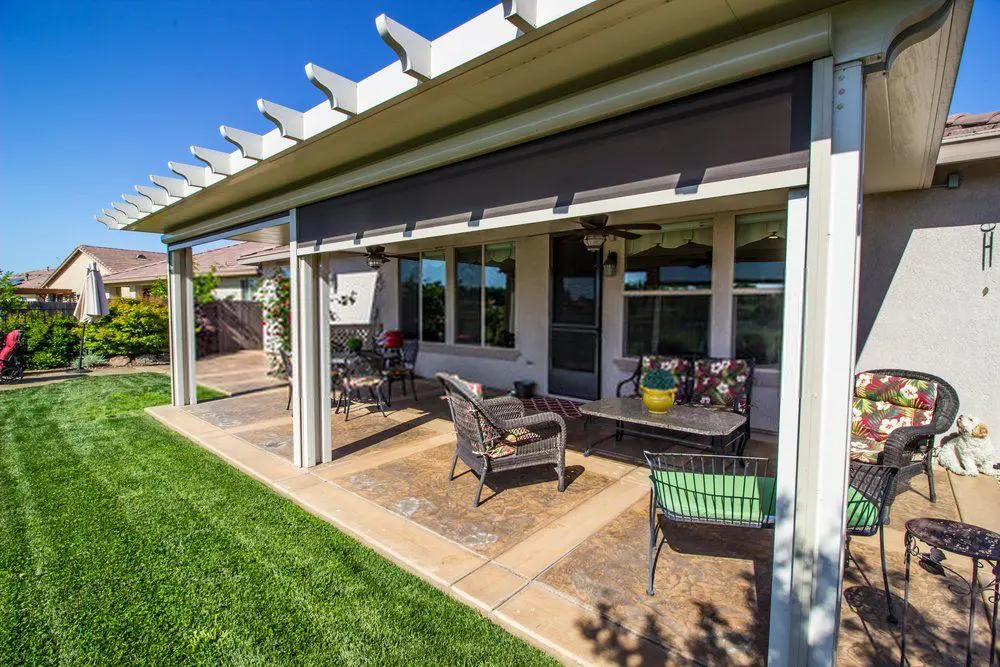 A covered patio with a trellis roof features outdoor furniture, including a glass-top table, chairs with patterned cushions, and a small potted plant on the table. The stamped concrete patio is adjacent to a well-manicured lawn and a house with large windows and a beige exterior.