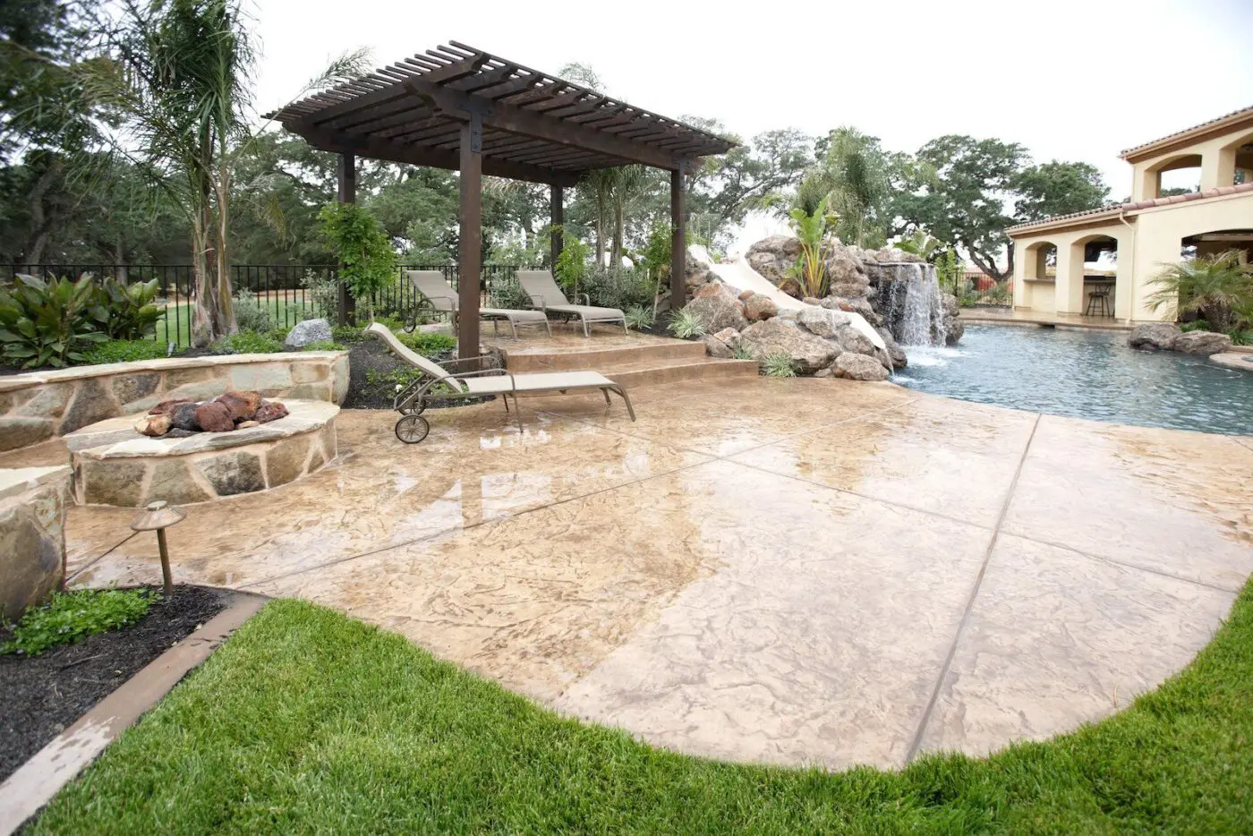A luxurious backyard in Reno featuring a swimming pool with a waterfall, two lounge chairs under a wooden pergola, and a stone fire pit. The stamped concrete pool decks complement the lush landscaping, offering a serene and elegant outdoor living space.