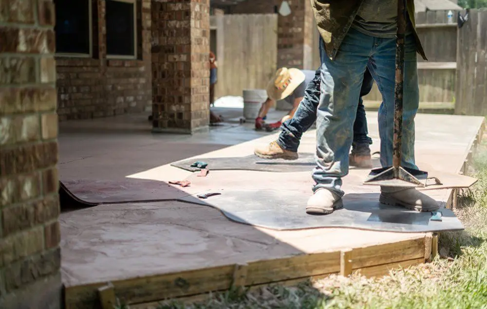 Construction workers laying a stamped concrete patio. One worker in boots is spreading concrete with a trowel while another worker in the background is smoothing out the surface. Tools and sheets of material are scattered on the ground around them, showcasing typical installation and repair processes in Reno and Sparks.