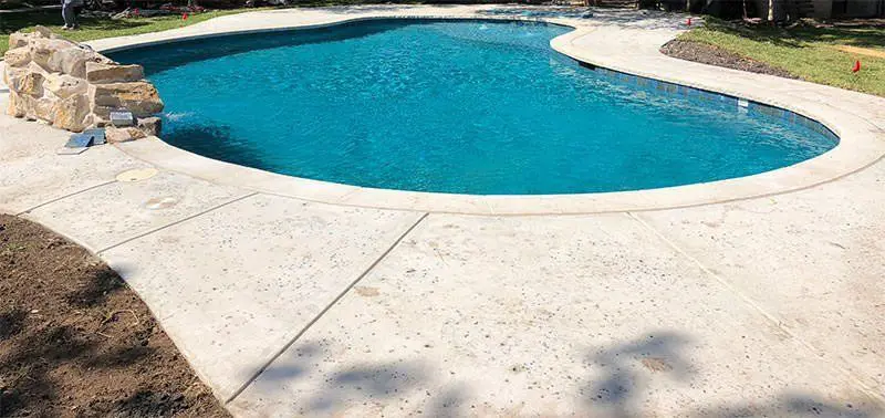 A serene backyard in Genoa NV features a clear blue, kidney-shaped swimming pool. The pool is surrounded by a smooth, light-colored deck installed by Reno Concrete Solutions, with a few stones and grass visible in the background, suggesting an inviting and tranquil outdoor space for relaxation and leisure.