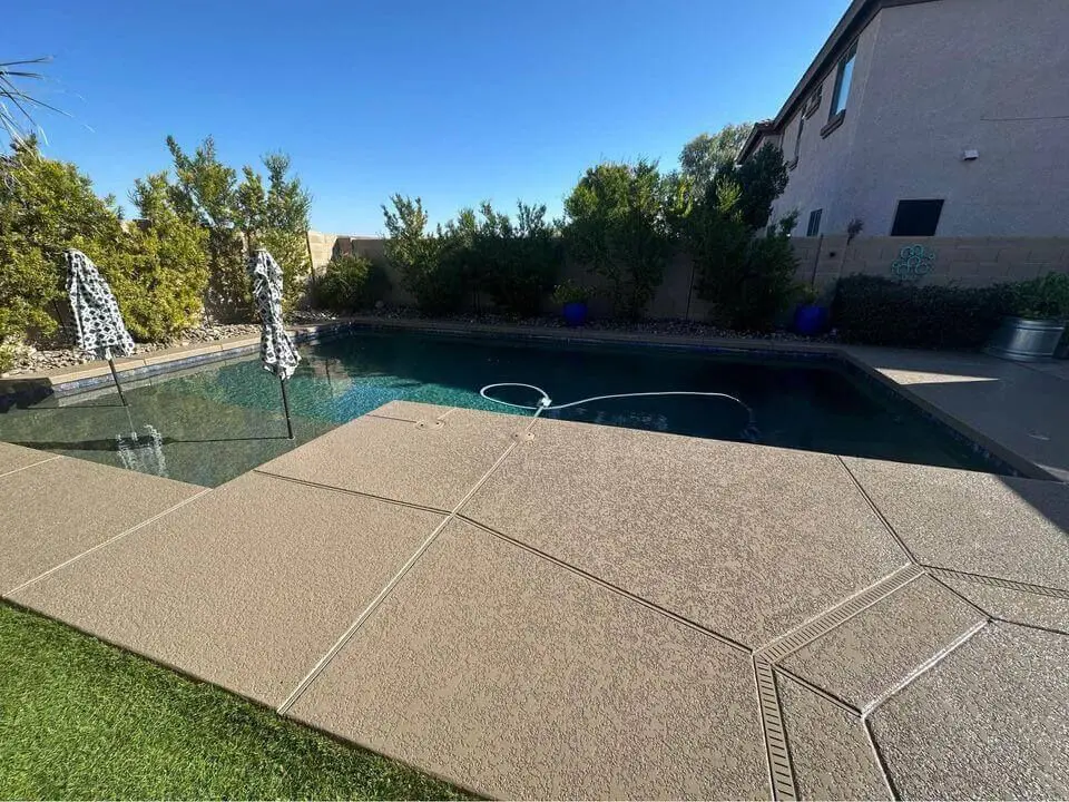 A backyard swimming pool with a clean, clear surface is partially shaded by patterned umbrellas. Surrounding the pool are shrubs and bushes providing privacy. The patio area is paved, showcasing the skill of concrete contractors in Carson City, NV. A neatly maintained grass patch rests at the edge, with a house visible in the background.