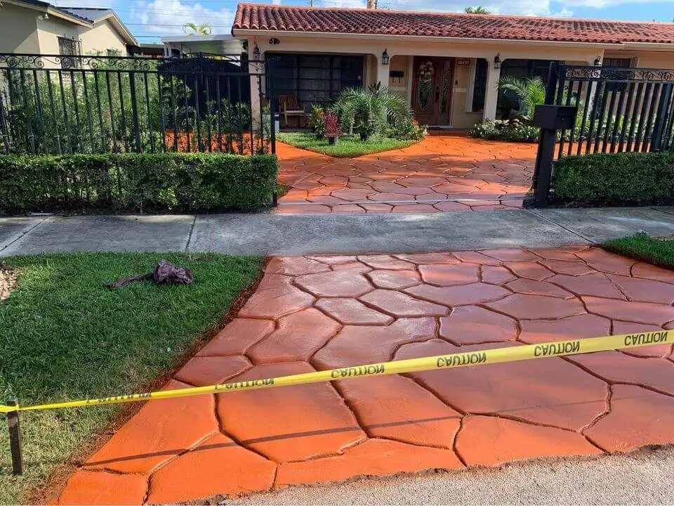 In Carson City, a residential project boasts a new orange-painted concrete driveway with a flagstone pattern. Yellow caution tape restricts access to the freshly painted area. A neatly trimmed green lawn flanks the driveway, and the house features beige walls and a red-tiled roof, thanks to skilled concrete contractors.