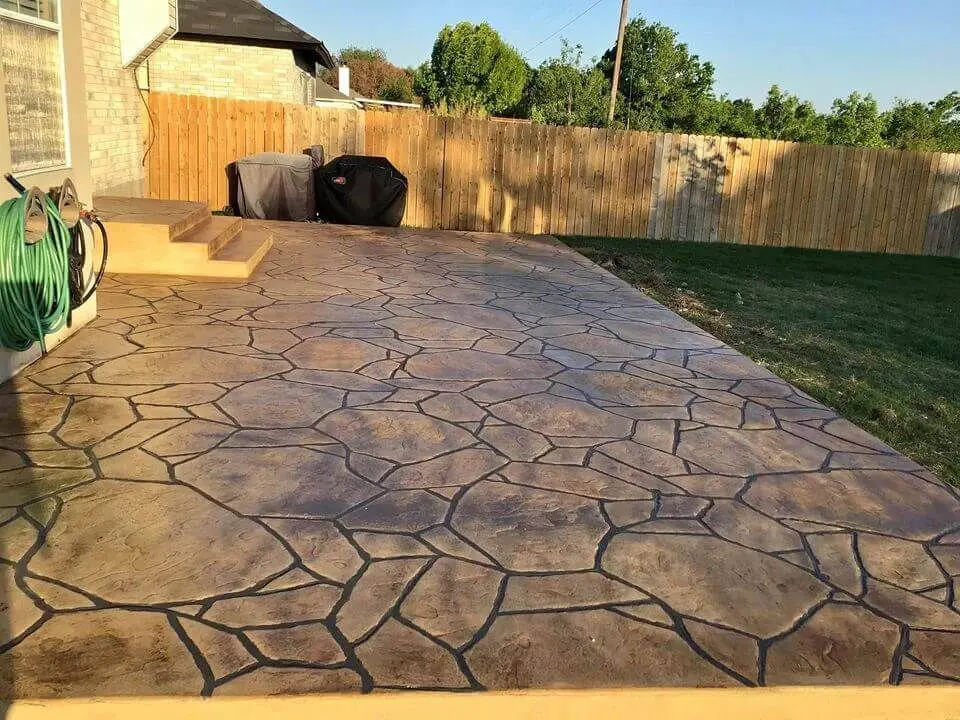 A backyard patio in Carson City NV with a decorative stone pattern, featuring a seamless blend of varying shades of brown. The patio is beside a small set of stairs leading to a house, with two black grills and a garden hose against the house's exterior wall. A wooden fence surrounds this beautiful residential project.