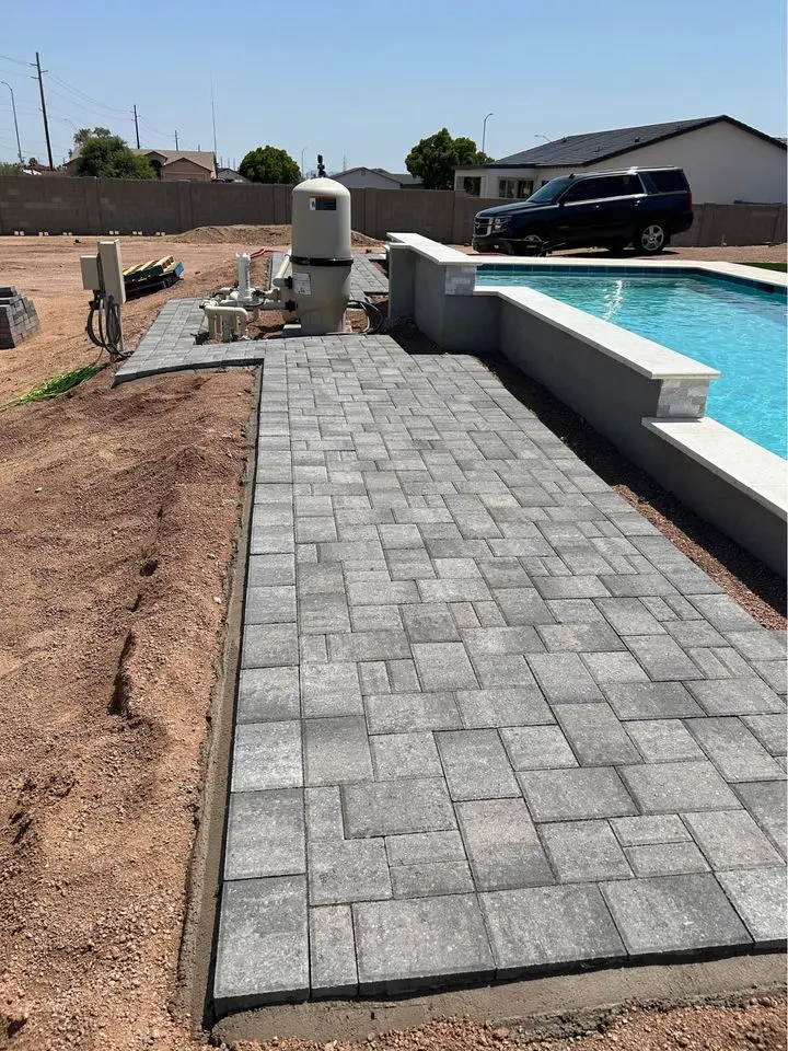 A backyard features a partially completed poolside patio with interlocking gray stone pavers. Nearby, construction equipment highlights ongoing paver installation, while a black SUV is parked beside the scene. The pool brims with water, reflecting the hustle of an area still under construction.