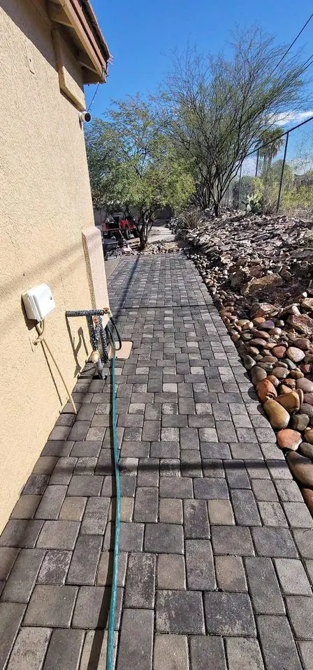 A long, paved pathway runs alongside a beige building, bordered by a rocky landscape with small trees. With expert pavers installation, the path extends like a driveway while a garden hose snakes along under the clear blue sky.