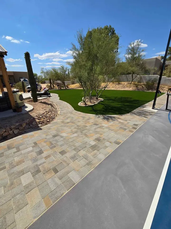 A backyard features a stone-paved pathway, showcasing expert pavers installation, leading to a lawn with desert plants and trees. To the right is part of a gray surface, possibly a pool or court. The landscape is under a clear blue sky with scattered clouds.