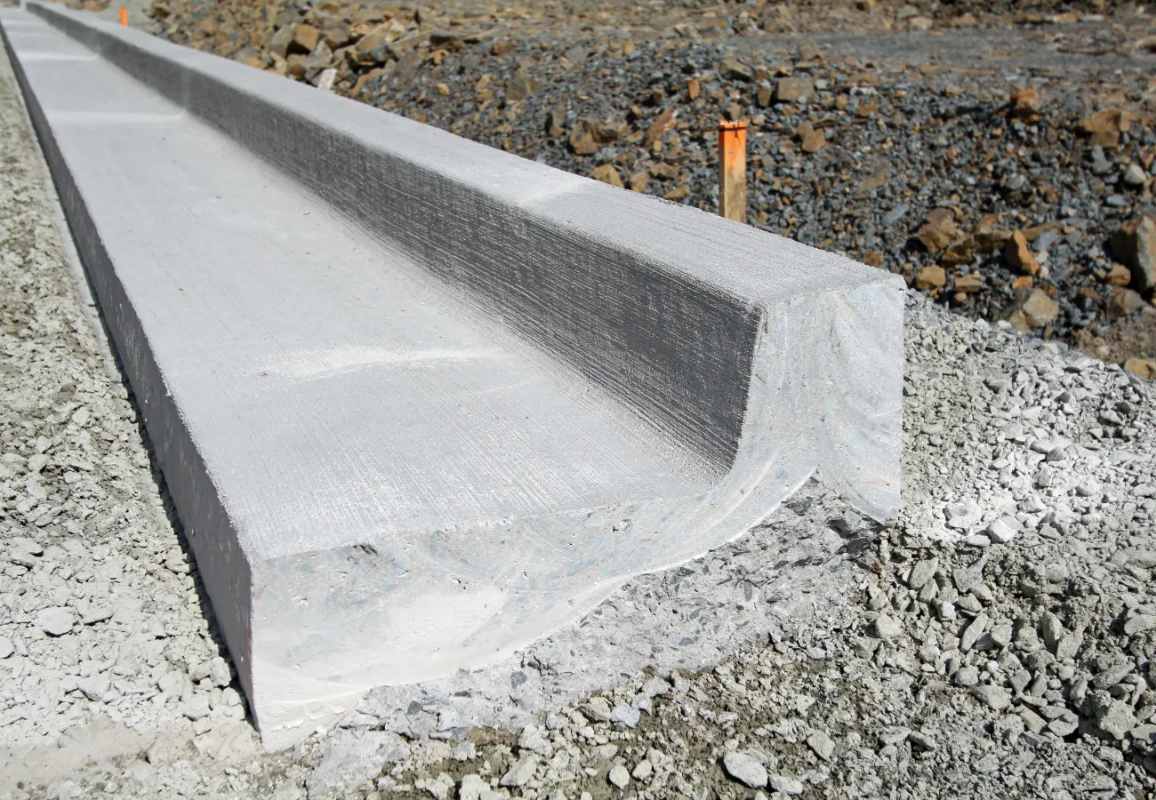 A close-up view of a freshly installed concrete curb amid a Carson City NV construction site, surrounded by gravel and rocky terrain. The curb's clean, smooth surface sits in stark contrast to the rough ground, with an orange marker stake in the background, showcasing expert work by local concrete contractors.