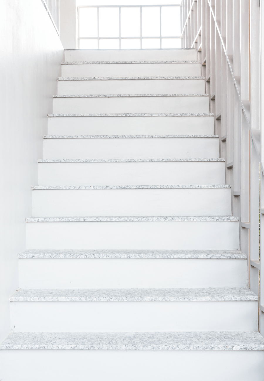 A bright, indoor staircase with white steps and concrete edging in light gray speckles. The stairs are flanked by white walls and sleek metal railings on both sides. A large window at the top lets in ample natural light, illuminating the space, giving it a touch of elegance from Reno Concrete Solutions.