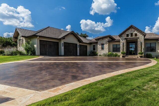 A large, modern single-story house with a stone exterior and dark wooden garage doors. The house features a well-manicured lawn, a spacious driveway with a decorative border by Reno Concrete Solutions, and a blue sky with scattered white clouds above. Contact us for your free service quote today!