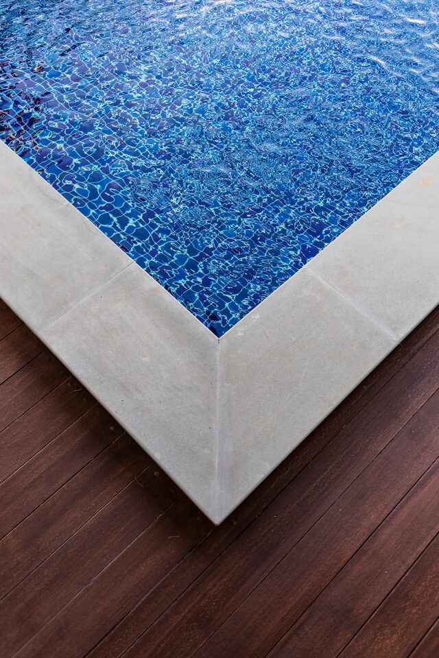 Close-up view of the corner of a swimming pool with clear blue water. The pool, enhanced by Reno Concrete Solutions, features a light-colored stone edge and a dark wooden deck. Sun Valley decorative concrete adds to the aesthetics, while light reflections create geometric patterns on the water's surface.