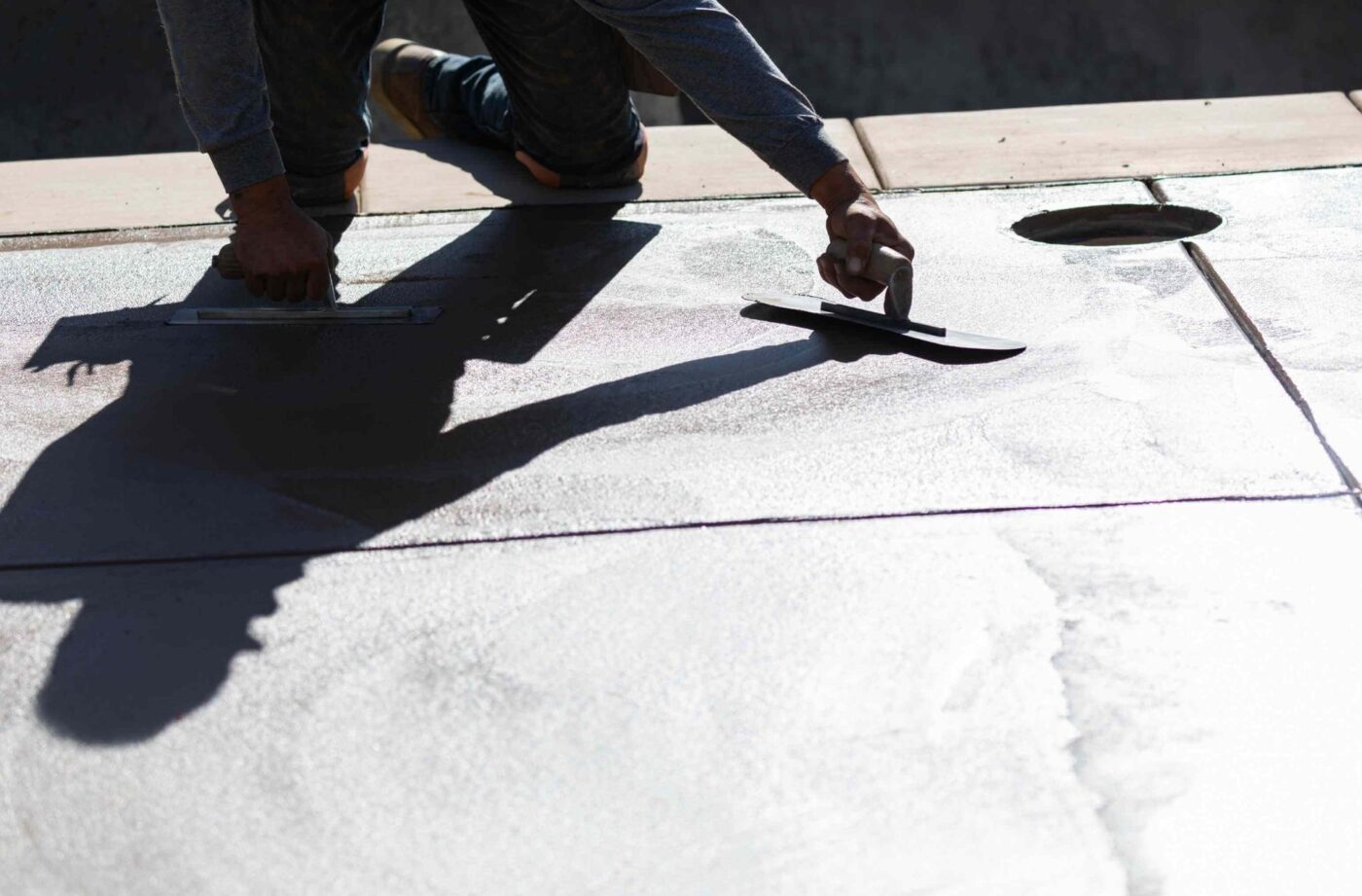 A licensed concrete contractor in Gardnerville NV is kneeling and smoothing a concrete surface with trowels, casting a shadow on the freshly dried cement. The sun is casting a strong light, highlighting the texture of the concrete. A circular opening is visible in the background—contact us for a free quote.