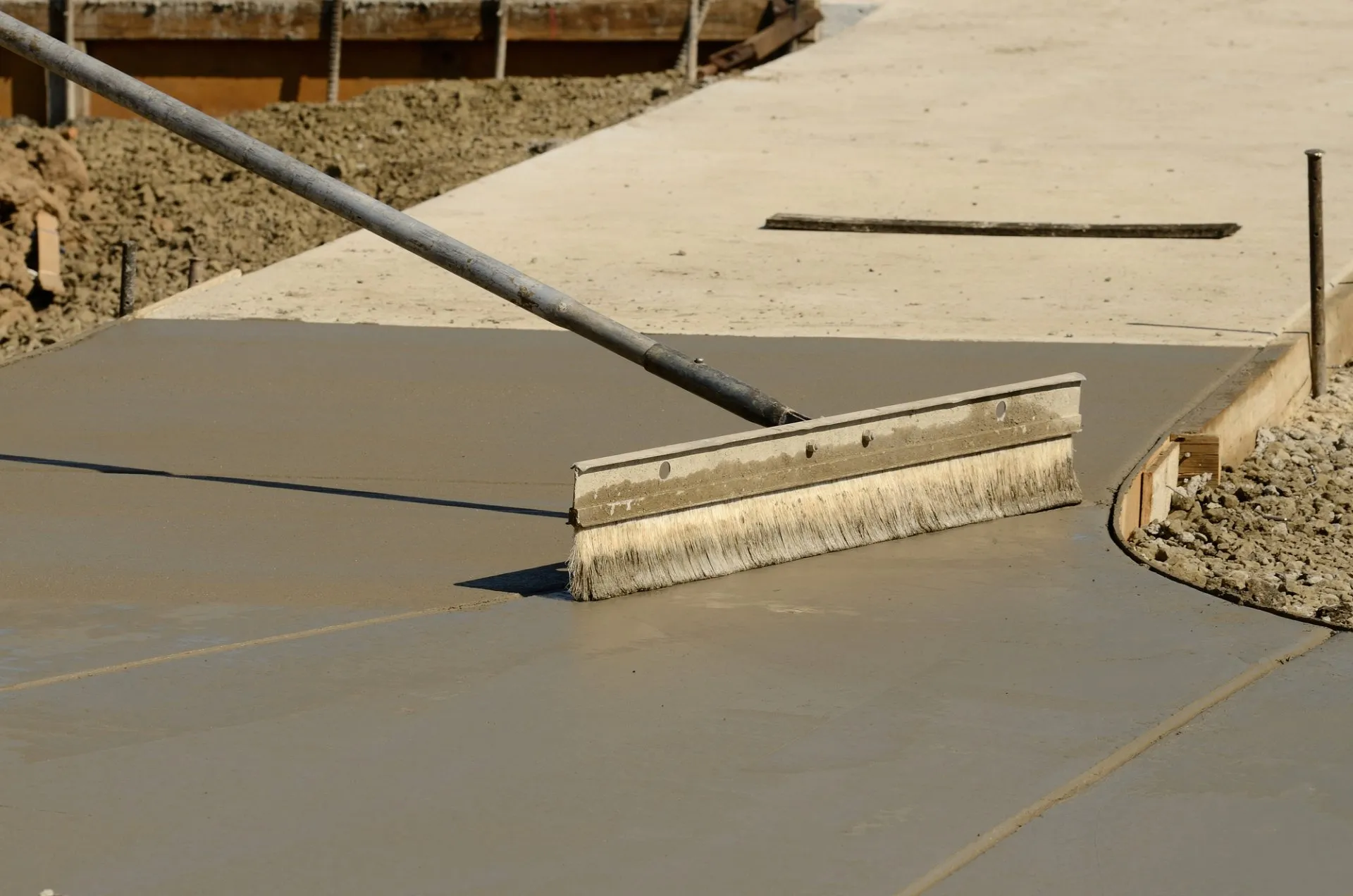 An industrial-grade broom is used to smooth the wet surface of a freshly poured concrete sidewalk at a construction site. The surrounding area shows dirt and other construction-related activities. For all your concrete needs, trust Reno Concrete Solutions for expert service and free quotes.